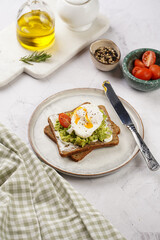 Trendy lifestyle sandwich: protein bread slice with cream cheese, mashed avocado, cherry tomatoes and poached egg on white scandi plate, light setting
