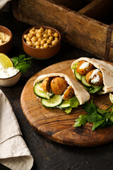 Traditional oriental chickpea deep fried falafel in flatbread pita, tzatziki yoghurt sauce, hummus, fresh lime and green cilantro on wooden board, dark surface