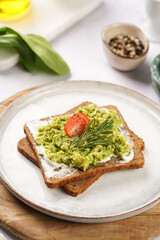 Fototapeta na wymiar Trendy lifestyle sandwich: protein bread slice with cream cheese, mashed avocado, cherry tomatoes and rosemary on white scandi plate, light setting