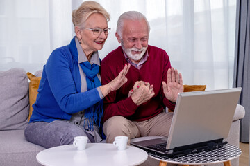 A lovely senior couple is sitting on a sofa and talking to their children and grandchildren via video call on a laptop in their living room. The elderly husband and wife are waving at a camera.