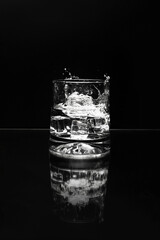 Glass with ice cubes and splash in clear liquid on black background with reflection