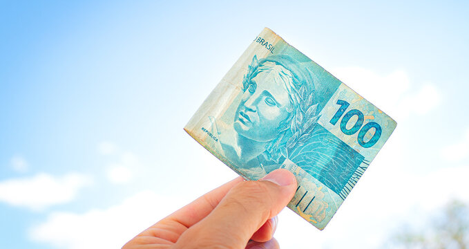 Um homem segurando com a m&atilde;o uma nota de 100 Reais. Fundo com c&eacute;u azul. Real Brasileiro - BRL, dinheiro do Brasil. Conceitos de economia brasileira e finan&ccedil;as.