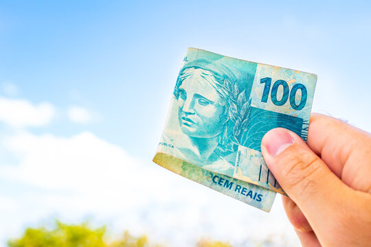 Real - moeda do Brasil. Uma c&eacute;dula do Real Brasileiro no valor de 100 Reais sendo segurada por um homem. Fundo com o azul do c&eacute;u e uma planta abaixo com algum espa&ccedil;o para c&oacute;pia. Economia brasileira.