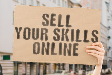 The phrase " Sell your skills online " on a banner in men's hand with blurred background. Ambition. Qualified. Tech. Gain. Return. Interest. Benefit. Work. Interest. Benefit. Wealth. Earn. Income