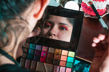 Beautiful latina woman looking in the mirror of her eye shadow palette, with her hand raised...