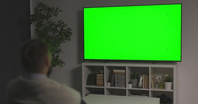 Back view of man watching green chroma key screen television while relaxing on couch. Male person spending time at home for screening of film.