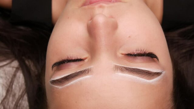 Master applies white markings to correct shape of eyebrows with a special brush. The beautiful face of young girl model on sofa in a beauty salon before microblading, permanent makeup eyebrow tattoo