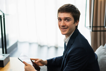 caucasian businessman smile, working with tablet, wear suit, sitting, looking at camera, copy space at window light background. Young man work business with home office background and copy space