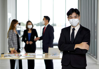 Young asian businessman in black suit fold his arms over the chest with confidence. The team is preparing for the meeting in the background. Everyone wears a face mask.