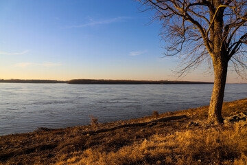 Fototapeta na wymiar a stunning shot of the blue running waters of the Mississippi river with bare trees and brown and yellow grass along the banks of the river with blue sky and clouds at sunset at Greenbelt Park 