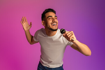 Portrait of cool young Arab man singing song, using microphone, performing karaoke in neon light