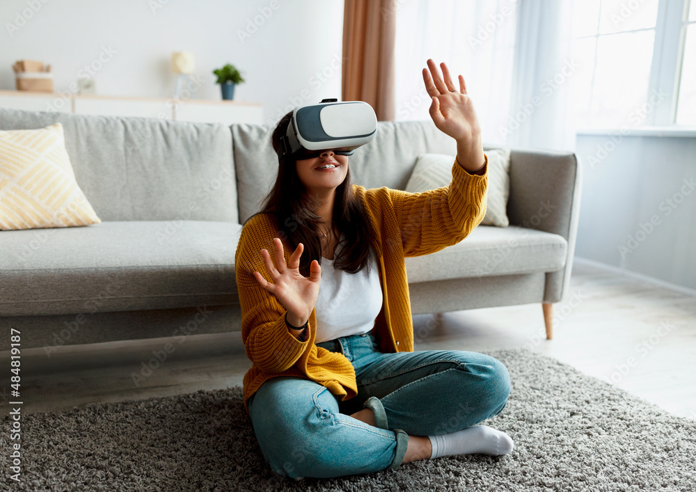 Wall mural Young middle eastern woman in virtual reality glasses exploring cyberspace, playing interactive video game at home