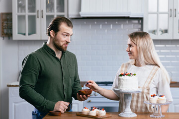 Pastry chef confectioner young caucasian woman with husband boyfriend man try taste of cake on kitchen table. Cakes cupcakes and sweet dessert