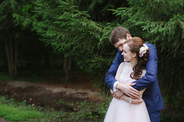 Naklejka premium the groom hugs the bride from behind against the background of spruce, they smile and look at each other with love