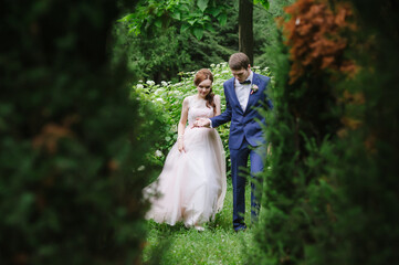 the bride and groom behind green trees and foliage walk holding hands and looking at their feet