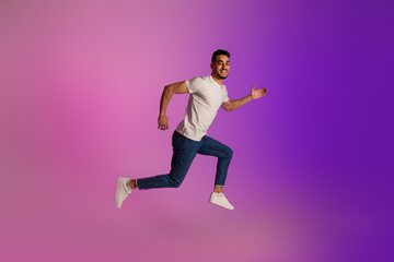 Fototapeta na wymiar Full length portrait of positive young Arab man in casual wear jumping in neon light, copy space