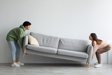 Happy young Asian couple moving sofa in living room, replacing furniture at home, copy space
