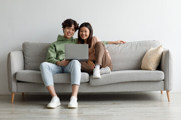 Happy young Asian couple using laptop, relaxing together on sofa at home, copy space