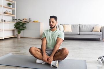 Fototapeta premium Peaceful millennial Arab guy sitting in lotus pose, meditating with closed eyes on mat at home, copy space