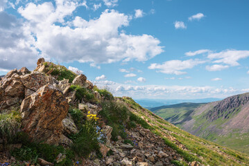 Scenic green landscape with sunlit pointy stone hill with grasses and flowers under clouds in blue sky. Colorful alpine scenery with sharp rock with grass and flowers in sunlight at changeable weather