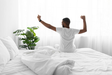 Fototapeta na wymiar Calm millennial african american guy sitting on bed stretching body and enjoying good morning