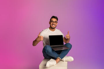 Young Arab guy holding laptop with empty screen, showing thumb up, recommending new website in neon light. Mockup - obrazy, fototapety, plakaty
