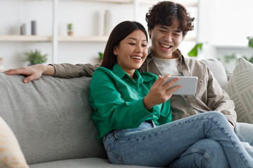 Obraz premium Millennial Asian couple using smartphone, relaxing on weekend, sitting on couch together at home, copy space