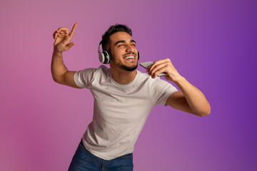 Young Arab man in headphones singing song, using smartphone as mic, enjoying new mobile music app...