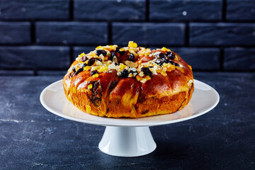 easter bread wreath sprinkled with candied orange, raisins and sliced peeled almonds on a white porcelain cake stand with a brick wall at the background