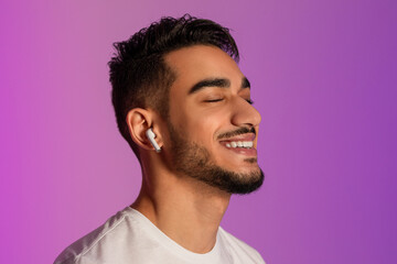 Obraz premium Handsome young Arab guy in casual outfit listening to music in earphones, closing eyes and relaxing in neon light