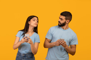 Fototapeta premium Privacy concept. Arab lady trying to look at her boyfriend's smartphone, guy hiding gadget, yellow studio background