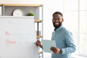 Smiling millennial african american bearded guy teacher in glasses stands near blackboard and teaches english