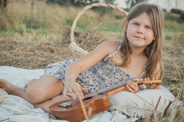 Portrait of smiling girl lying on blanket in dry hay field, having picnic, learning playing guitar...