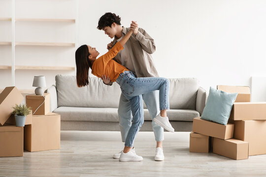 Affectionate young Asian couple dancing in their house among cardboard boxes on moving day, full length