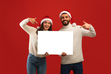 Happy arab couple in red Santa hats holding white empty board for advertisement, pointing on...