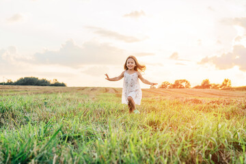 Fototapeta na wymiar Portrait of smiling girl playing, jumping and running on grass hay field paths of dry grass in the sunset. Waving hands. Forest on bright light background. Cloudy sunny sky. Haying time