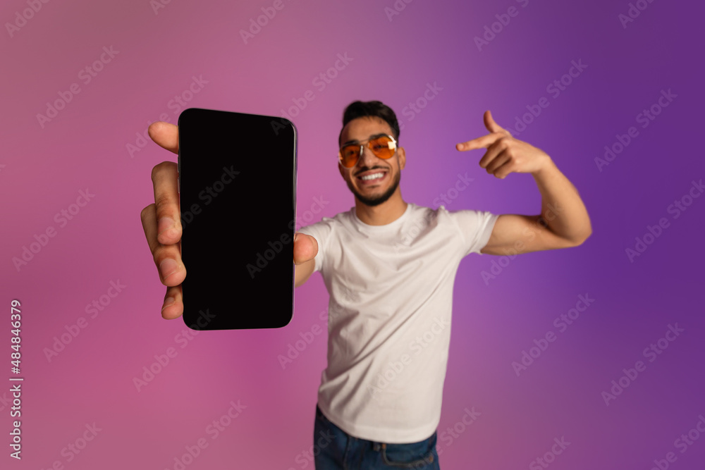 Poster cool young arab guy pointing at cellphone with empty black screen, recommending new app or website i