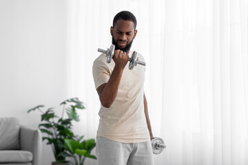 Strong young black male with beard in white sportswear raises dumbbell in room interior on window background