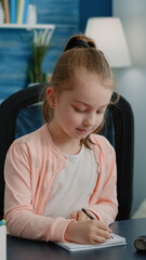 Schoolgirl writing on notebook with pen for homework. Little child concentrated on task for online remote lessons while sitting at desk with computer and textbook. Homeschooled pupil