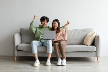 Young Asian couple with laptop gesturing YES, celebrating online business success, sitting on couch...