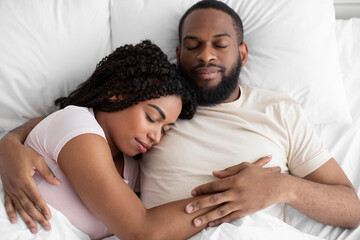 Calm young african american guy and woman sleeping, hugging on white bed in bedroom, top view