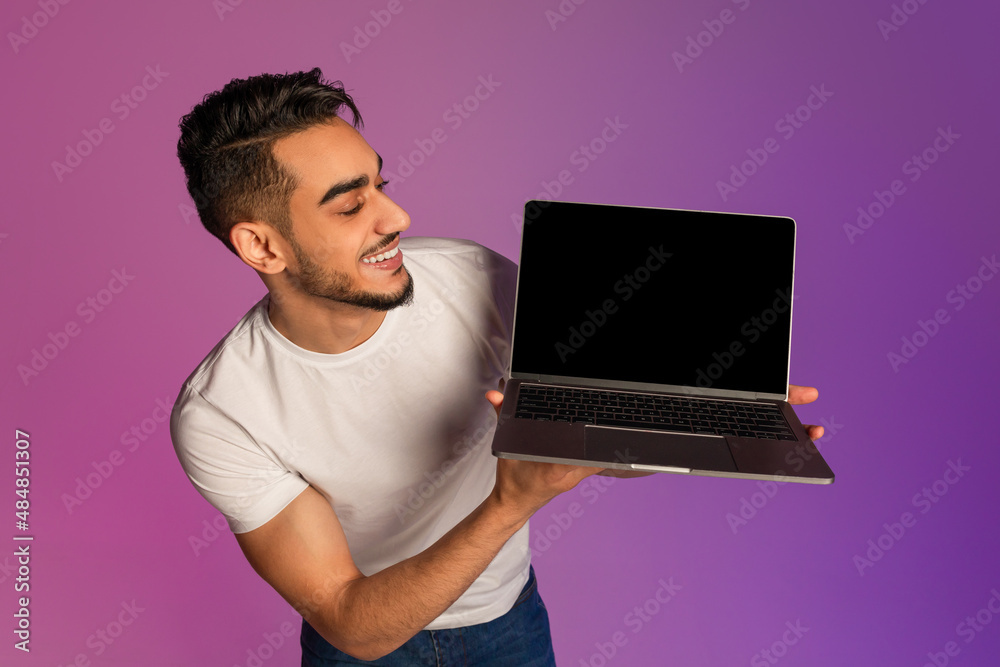 Poster millennial arab man showing laptop computer with empty black screen in neon light, mockup for new we