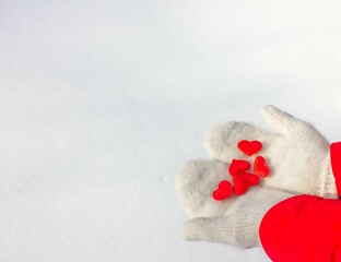 Fototapeta premium Female hands in knitted white mittens hold a silk red heart on a snowy background. Concept of love and valentine.
