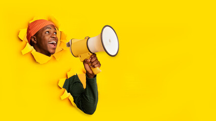 Emotional young black guy shouting into megaphone through hole in ripped orange paper, banner with free space