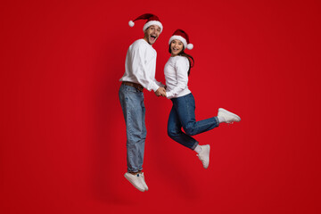 Overjoyed Couple Wearing Santa Hats Holding Hands And Jumping Over Red Background