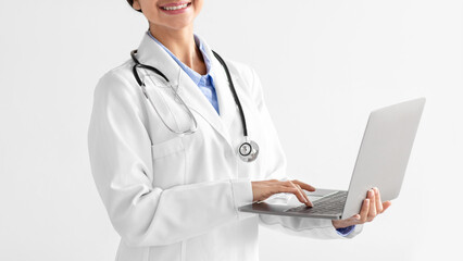 Cropped young indian lady doctor in white coat with stethoscope typing on computer on light...