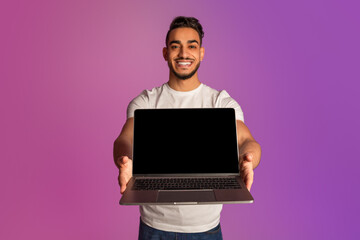Happy millennial Arab man holding laptop with black blank screen, showing free space for your mockup in neon light