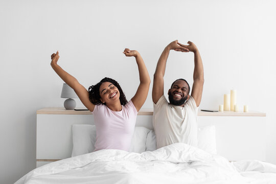 Happy millennial black husband and wife woke up, stretching bodies on white bed