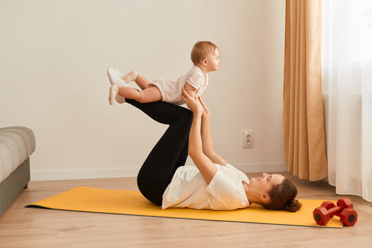 Side view of a woman in black leggings and a white T-shirt, a girl lying on the floor on a mat on her back with her legs raised up and holding a small child, home coach.