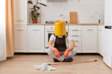 Portrait of unknown anonymous male housekeeper sitting on floor in kitchen with yellow bucket on his head, cleaning house, washing floor, being tired.